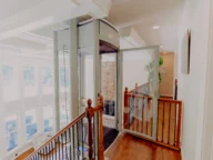 Modern home with a glass-enclosed elevator on the second floor landing, featuring wooden railings and large windows in the background.