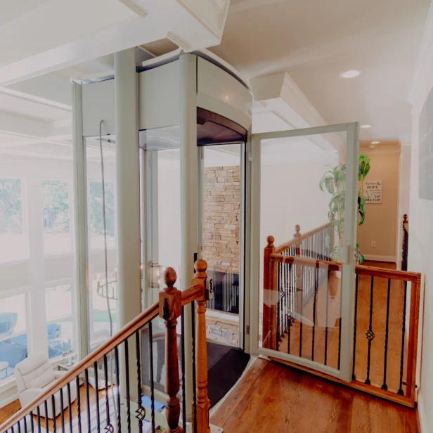 Modern home with a glass-enclosed elevator on the second floor landing, featuring wooden railings and large windows in the background.