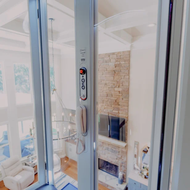 A glass elevator with a phone and control panel inside, overlooking a modern living room with large windows and a brick fireplace.