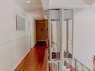 A home interior featuring a second-floor hallway with wooden flooring, leading to multiple rooms. A glass elevator is adjacent to the staircase with wooden railings, descending to the ground floor.