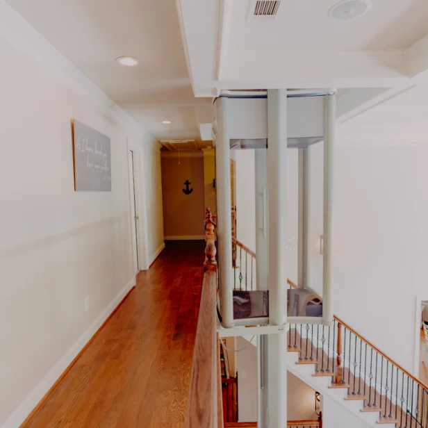 A home interior featuring a second-floor hallway with wooden flooring, leading to multiple rooms. A glass elevator is adjacent to the staircase with wooden railings, descending to the ground floor.