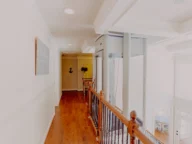 A well-lit hallway with wooden floors and a wooden railing overlooking a living area. An elevator and an anchor decoration are visible at the end of the hallway.