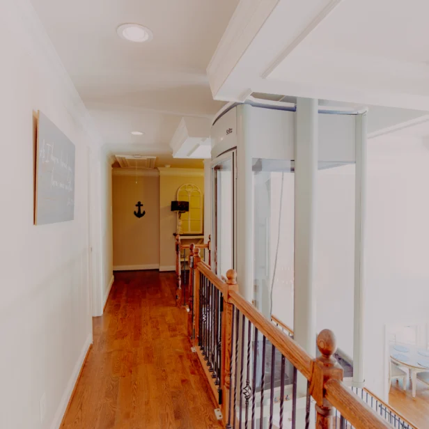 A well-lit hallway with wooden floors and a wooden railing overlooking a living area. An elevator and an anchor decoration are visible at the end of the hallway.