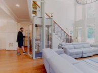 A spacious living room with high ceilings features a man operating a small glass elevator. The room includes modern gray sofas, large windows, and a wooden staircase.