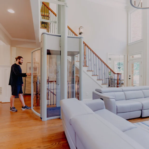 A spacious living room with high ceilings features a man operating a small glass elevator. The room includes modern gray sofas, large windows, and a wooden staircase.