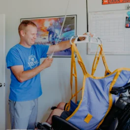 A man in a blue shirt operates an overhead patient lift to assist a person in a wheelchair. A calendar and a Star Wars poster are on the wall behind them.