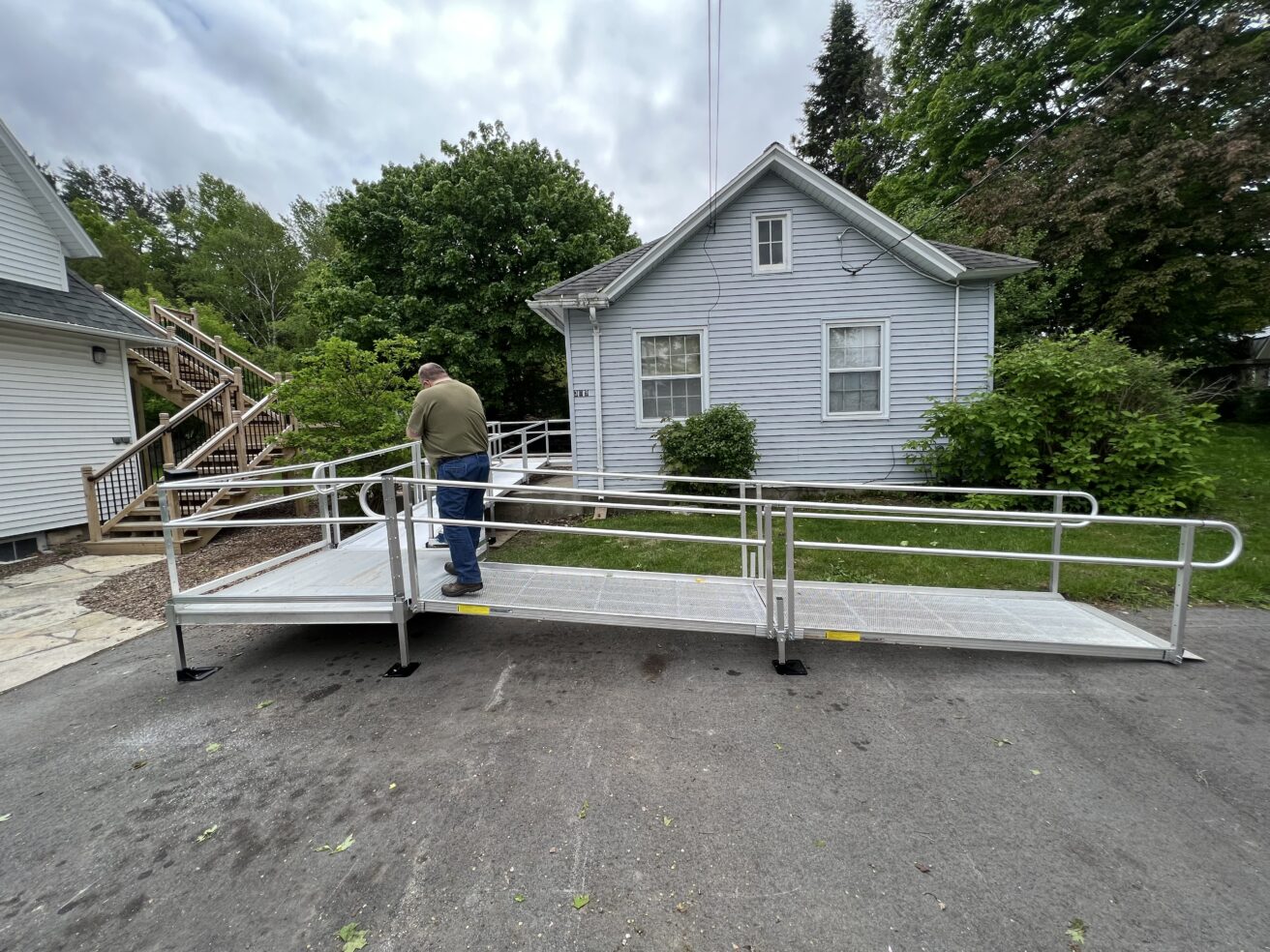 A man stands on a metal accessibility ramp leading to the entrance of a small white house surrounded by trees and shrubs.