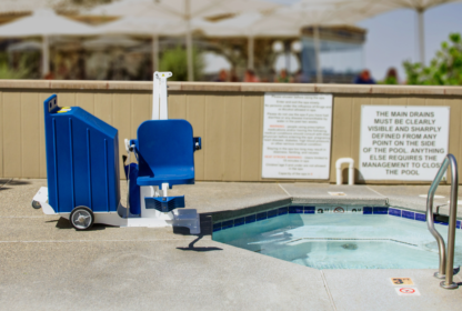 A blue pool lift is positioned next to a small, tiled whirlpool or spa on a concrete patio, with safety signs visible in the background.