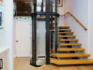 Modern interior with a glass elevator and a wooden floating staircase adjacent to a white door.