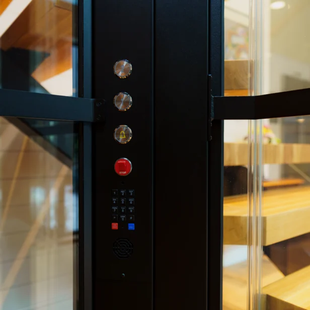 Close-up of a modern elevator control panel with four buttons, including a red emergency button, and a keypad inside a transparent elevator with wooden stairs visible in the background.