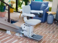 A stair lift chair installed on a staircase in a living room. The chair features a cushioned seat, armrests, and footrest, designed for mobility assistance. Plants and furniture are in the background.