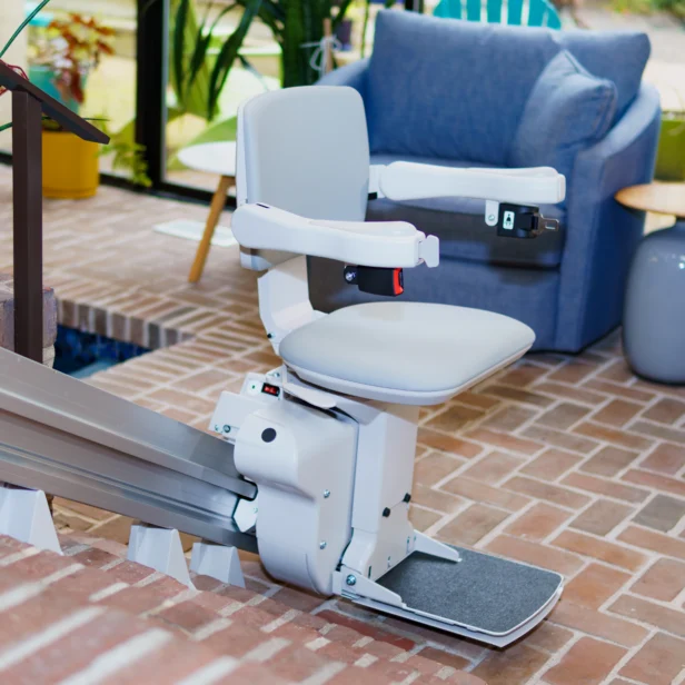 A stair lift chair installed on a staircase in a living room. The chair features a cushioned seat, armrests, and footrest, designed for mobility assistance. Plants and furniture are in the background.