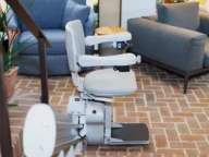 A stairlift installed in a living room setting, with a grey seat and armrests, shown on a brick floor next to sofas and plants.