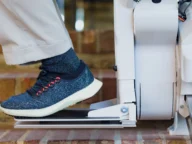 A person wearing blue sneakers and beige pants is using a stairlift, descending a brick staircase.