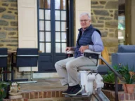 An elderly man smiles while seated on a stairlift, positioned on a staircase in a cozy home setting. In the background, a dining area and potted plants add to the warmth of the space.