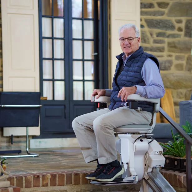 An elderly man smiles while seated on a stairlift, positioned on a staircase in a cozy home setting. In the background, a dining area and potted plants add to the warmth of the space.