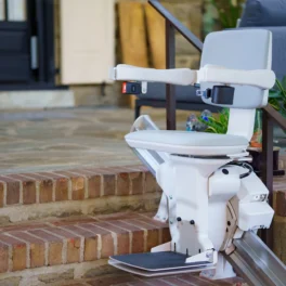 A white stairlift installed on a residential staircase with brick steps, leading up to a dark-colored front door. Plant pots and furniture are visible in the background.