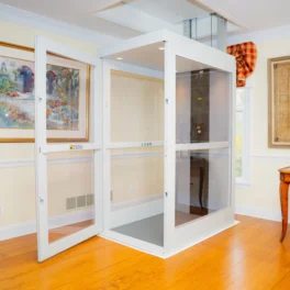 A residential indoor elevator with open glass doors, situated in a well-decorated room with paintings on the walls and wooden furniture.