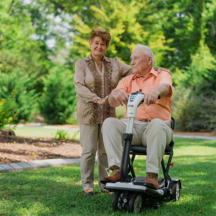 An elderly man sits on a mobility scooter while an elderly woman stands beside him, both in a garden setting with greenery and a small shed in the background.