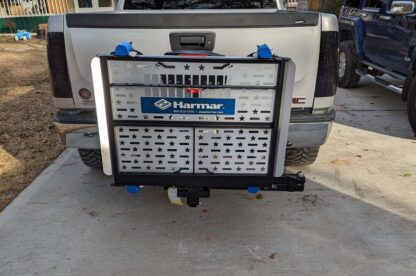 A white GMC pickup truck with a Harmar lift platform attached to its rear, parked on a concrete driveway in front of a wooden house.