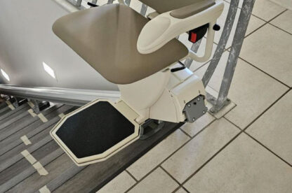 A beige stair lift chair installed on a staircase with a metal railing, ready for use to assist individuals in moving up and down the stairs.