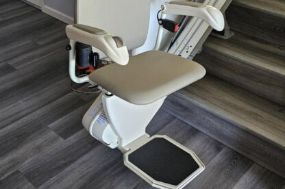 A beige stairlift chair installed adjacent to a staircase in a modern home with gray wood flooring.