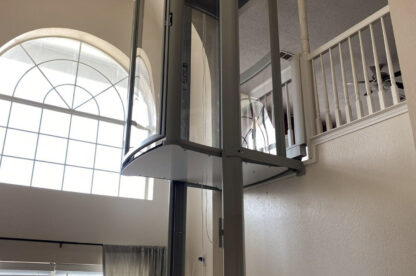 A residential elevator installed inside a double-height living room, with large arched windows and a loft area visible above.