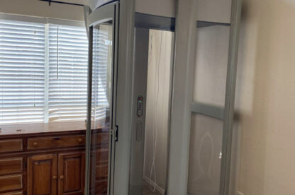 A home elevator with glass doors and a gray frame is installed next to wooden cabinets in a room with tiled flooring and a large window.