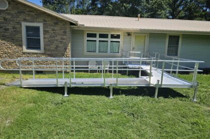 A metal wheelchair ramp leads to the front door of a house with stone and siding exterior, set on a grassy lawn with trees in the background.