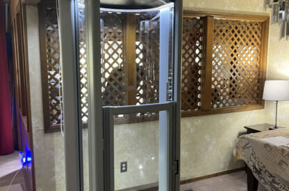 A compact, glass-enclosed residential elevator installed in a home with beige walls, wooden lattice paneling, and carpeted floors.