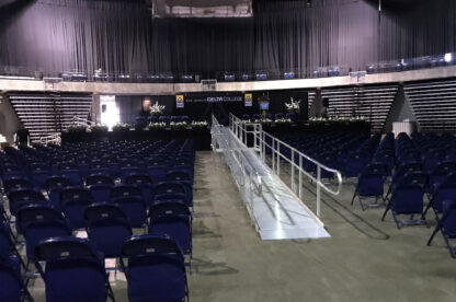 An empty auditorium set up for a graduation ceremony with rows of blue chairs facing a stage adorned with flowers and a "Delta College" banner.