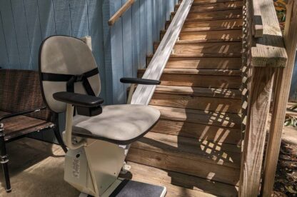 A stairlift installed alongside a wooden outdoor staircase, with a padded chair and footrest on a metal rail, providing mobility assistance.