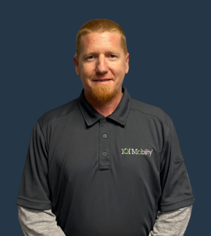 A man with short red hair and a goatee, wearing a gray "IOU Mobility" polo shirt stands against a solid dark background.