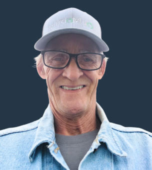 Smiling older man with glasses wearing a gray cap and a denim jacket against a dark background.