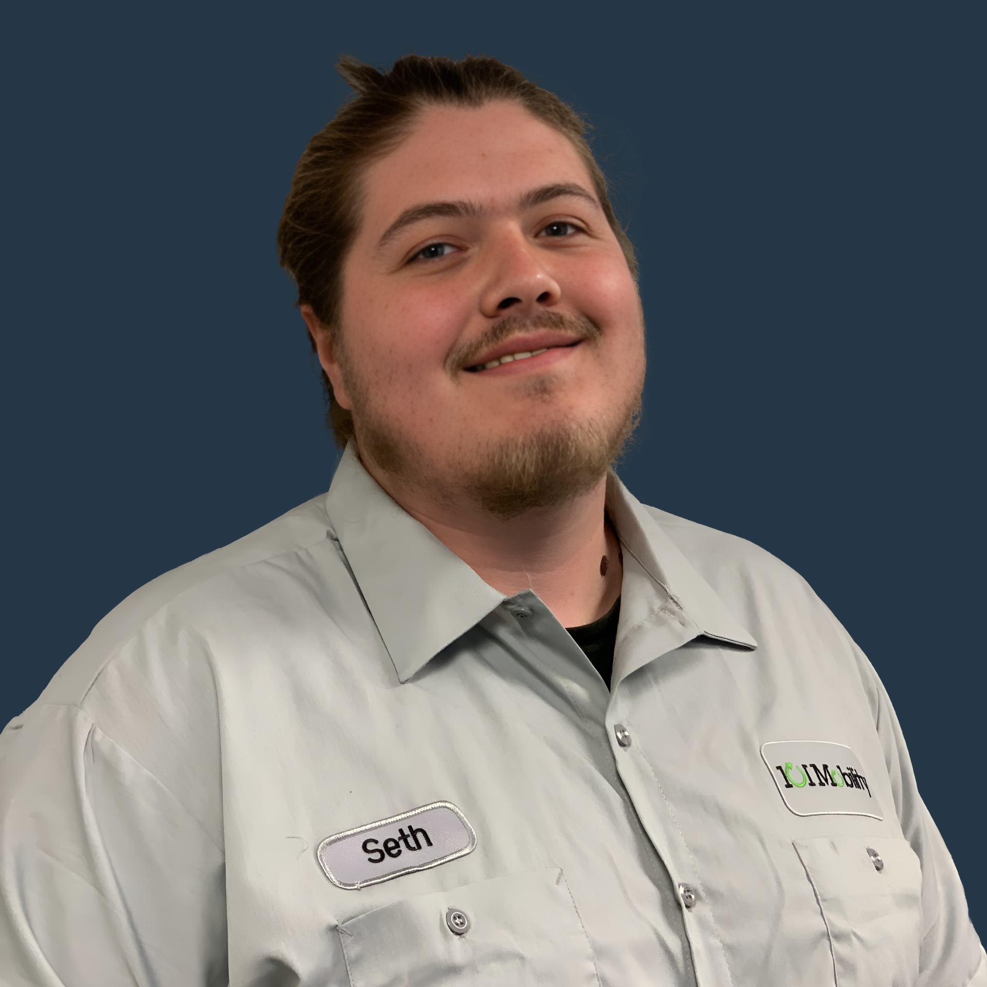 A person with a beard and mustache, smiling, wearing a light gray work shirt with a name tag that reads "Seth" and a logo on the other side against a dark blue background.