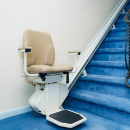 A beige stairlift is installed next to a flight of blue carpeted stairs. The seat has armrests and a footrest, and it is positioned at the bottom of the staircase.