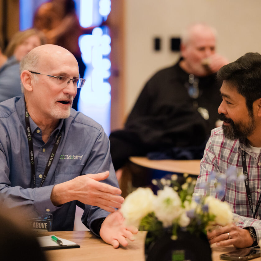 Two men are seated at a table engaged in conversation at a casual event. Flowers and papers are on the table, and other people are visible in the background.