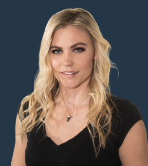 A woman with long blonde hair and light makeup is wearing a black top and a shark tooth necklace, standing against a solid dark blue background.