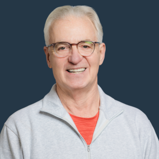 A smiling elderly man with white hair and glasses wearing a light gray zip-up jacket over an orange shirt stands in front of a dark blue background.