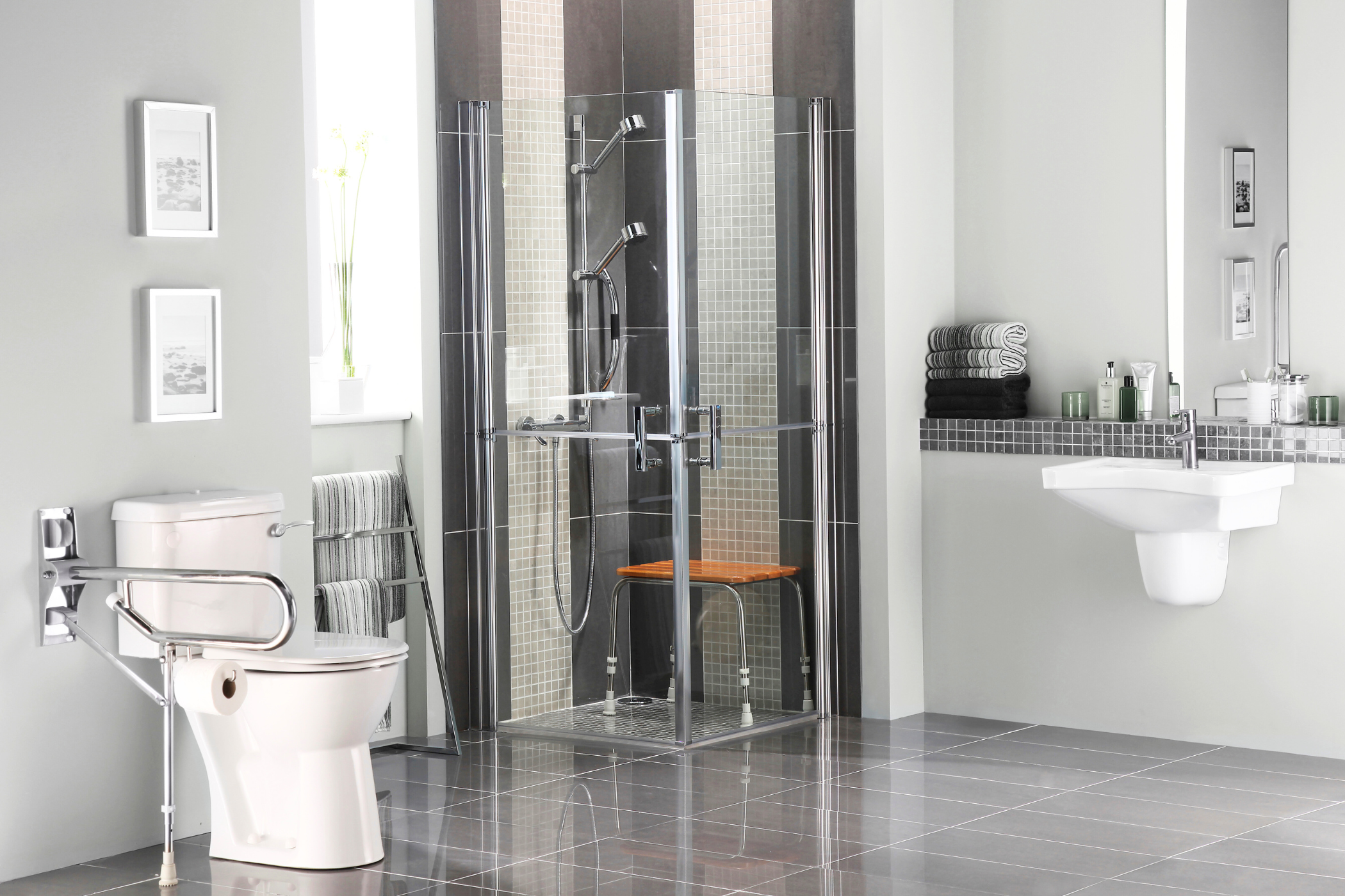 Modern bathroom with a glass-enclosed shower, a white toilet with a grab bar, a wall-mounted sink, and folded towels on a shelf. The floor is tiled, and there are framed pictures on the wall.