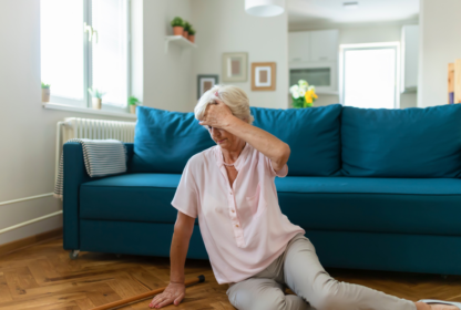 An elderly woman sits on the floor in front of a blue couch, holding her head with one hand and a walking cane lying beside her. The living room is brightly lit and furnished.