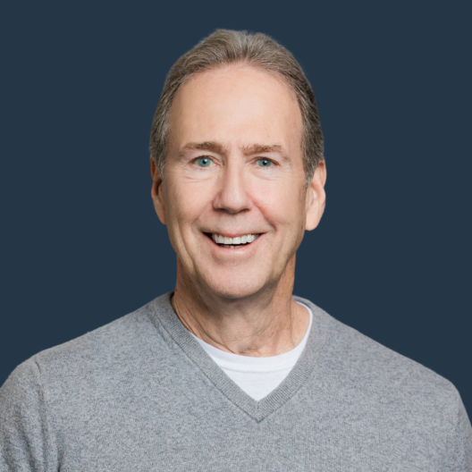 A man with gray hair and blue eyes, wearing a gray sweater and white shirt, smiles at the camera against a plain dark blue background.