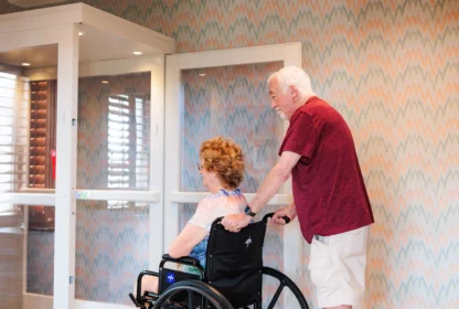 An elderly man pushes a woman in a wheelchair in a brightly lit hallway with patterned wallpaper.