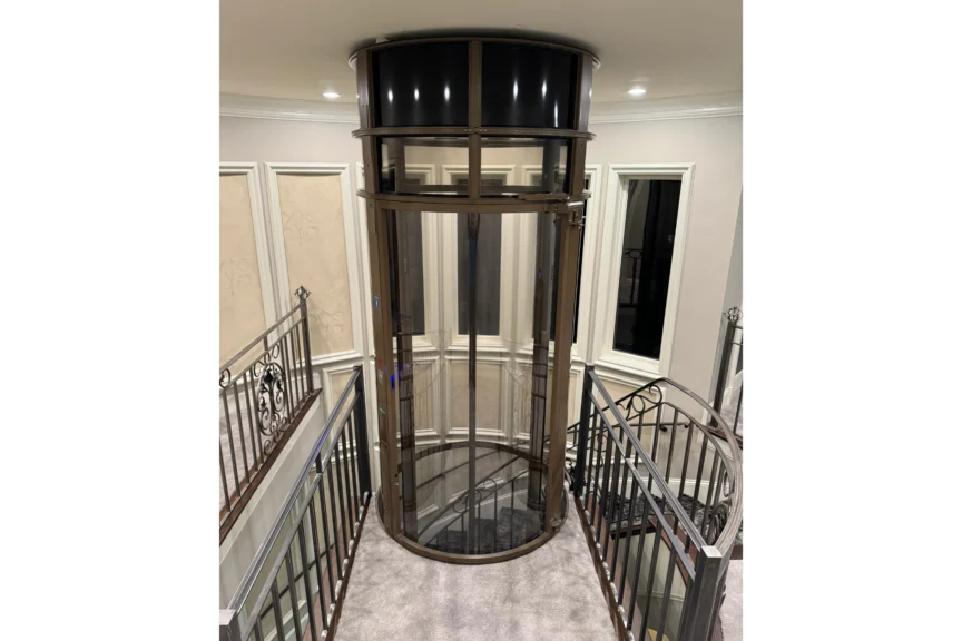 A modern glass elevator with a cylindrical shape is situated between ornate metal railings and centered within a room with beige walls and recessed lighting.