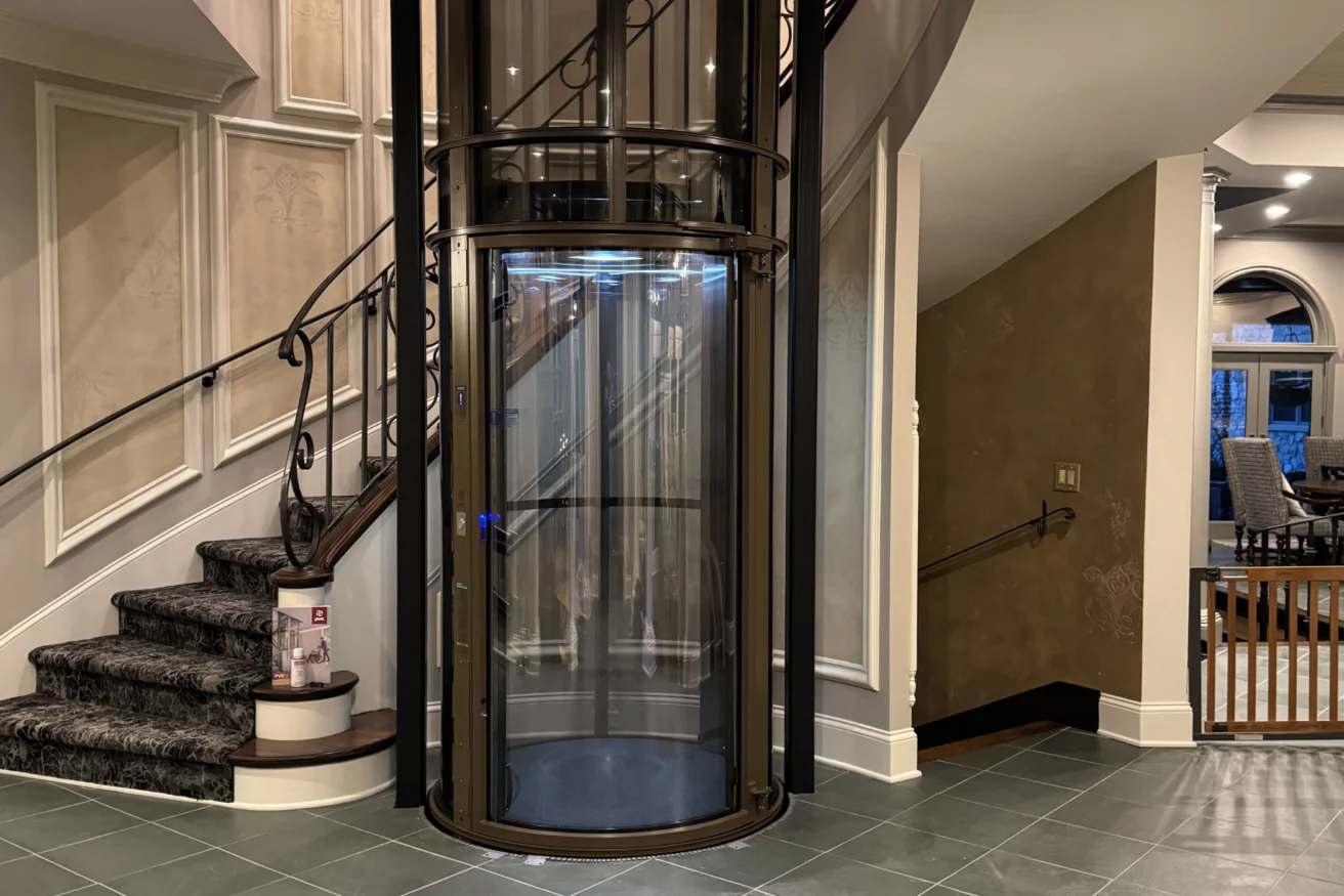 A glass cylinder elevator next to a curved staircase in an elegant foyer with tiled floors and decorative walls.