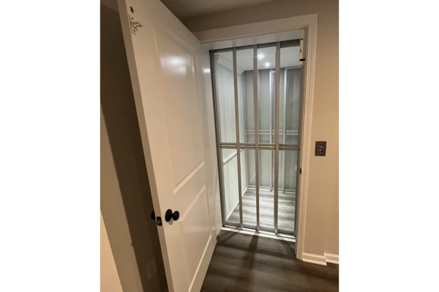 A small residential elevator with a metal gate is located inside a room, with the door open to show the interior. The floor is wood-paneled.