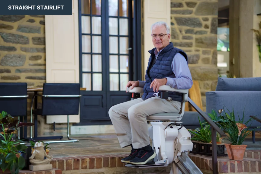 An older adult seated on a stairlift in a home setting, with stone walls and a dining area visible in the background.