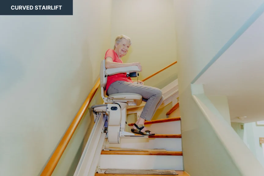 An older adult smiling while using a curved stairlift on a staircase in a home.