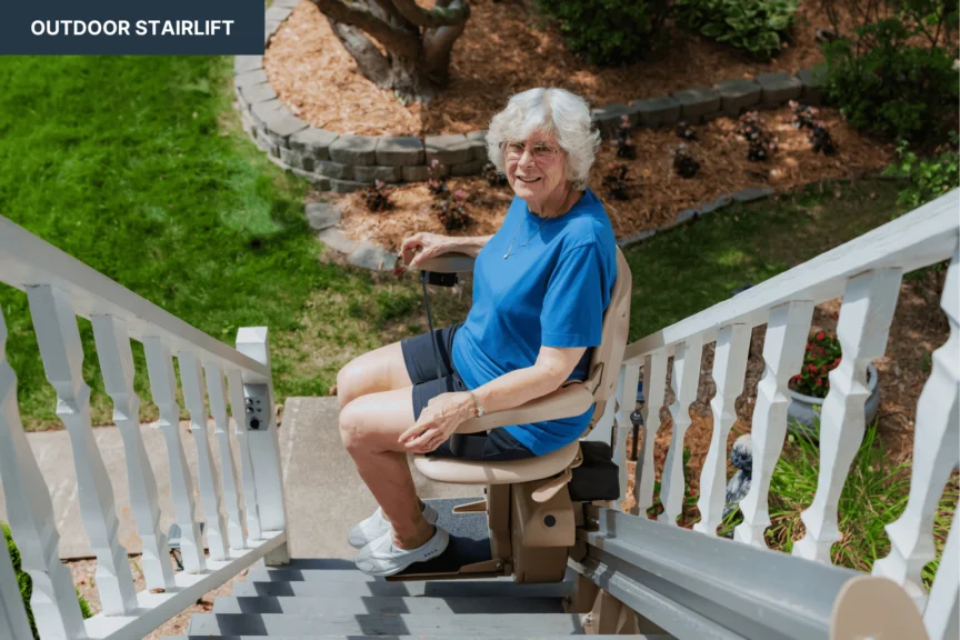 An older woman sits on an outdoor stairlift, smiling. She is wearing a blue shirt and shorts, with a garden visible in the background.