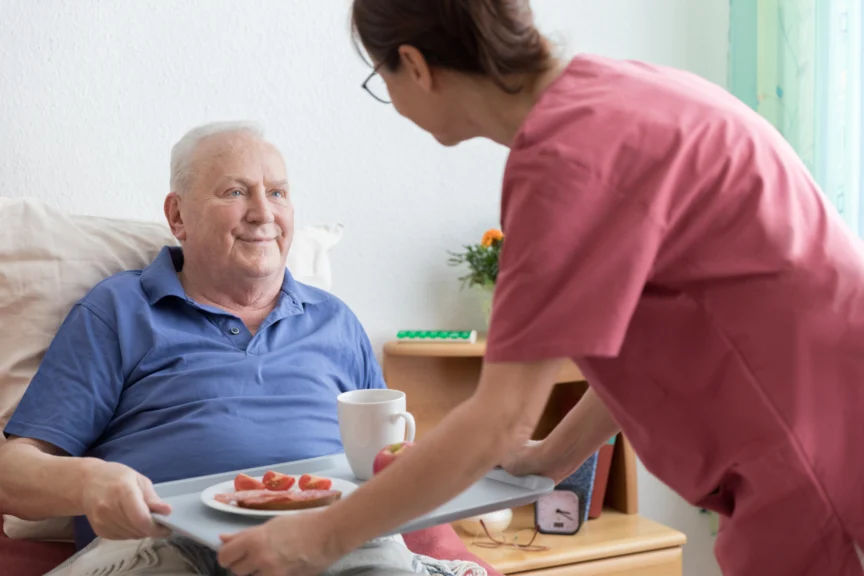 A caregiver serves breakfast to an elderly man sitting in bed, including a tray with a mug and a plate of food.